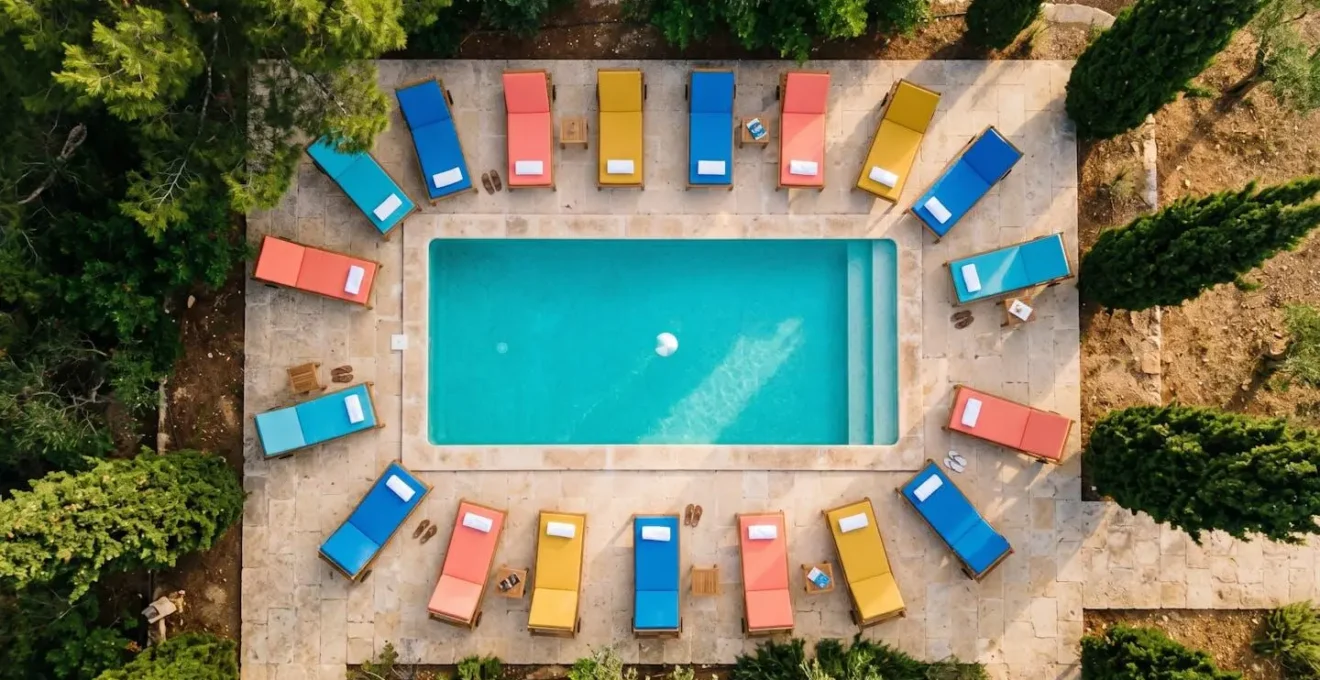 Vue aérienne d'une piscine extérieure entourée d'un cercle de chaises longues colorées avec un ballon au centre, bordée de pins maritimes sous lumière dorée matinale